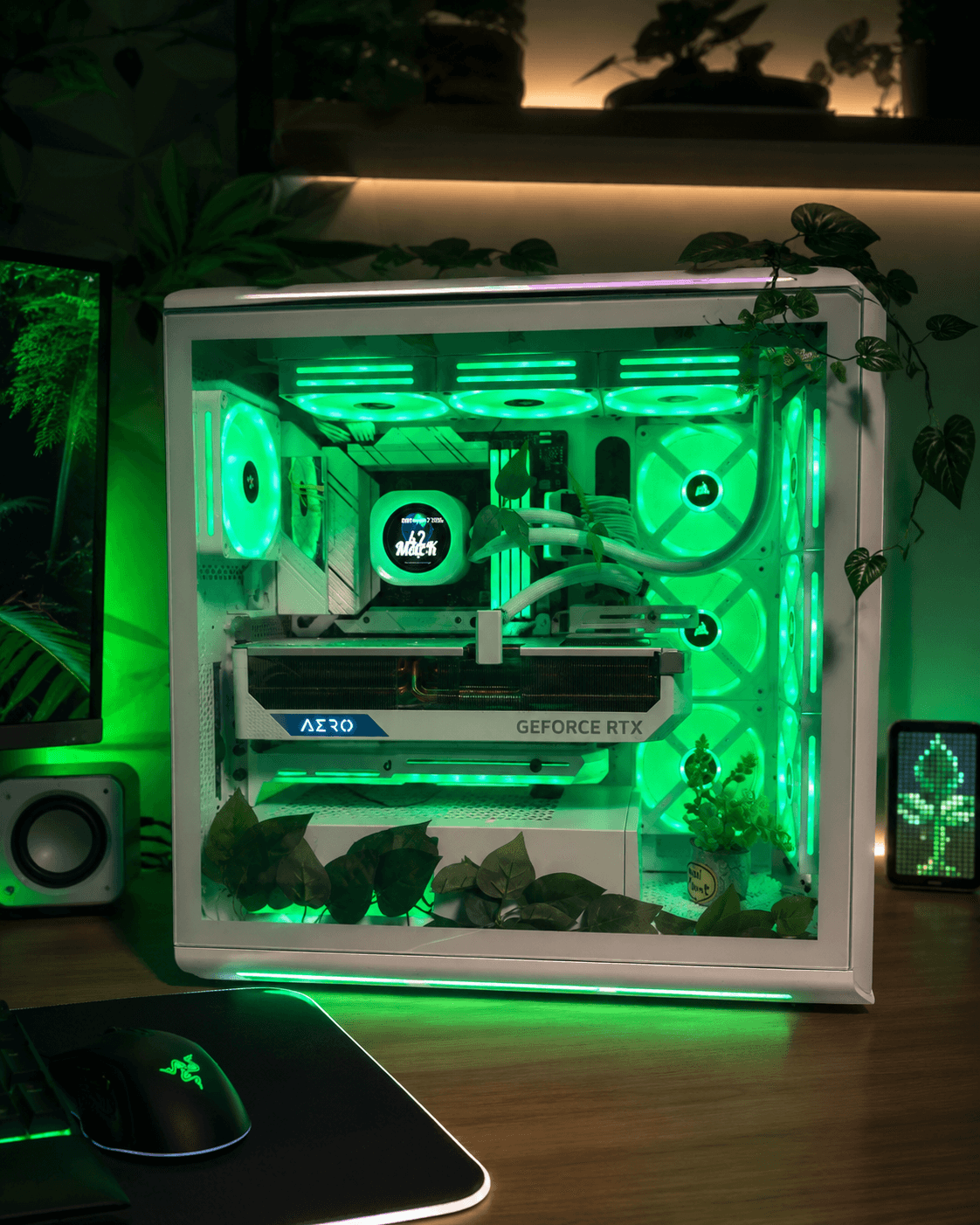 White PC case with a clear side panel and green internal lighting, showing a GeForce RTX graphics card, on a desk surrounded by plants.